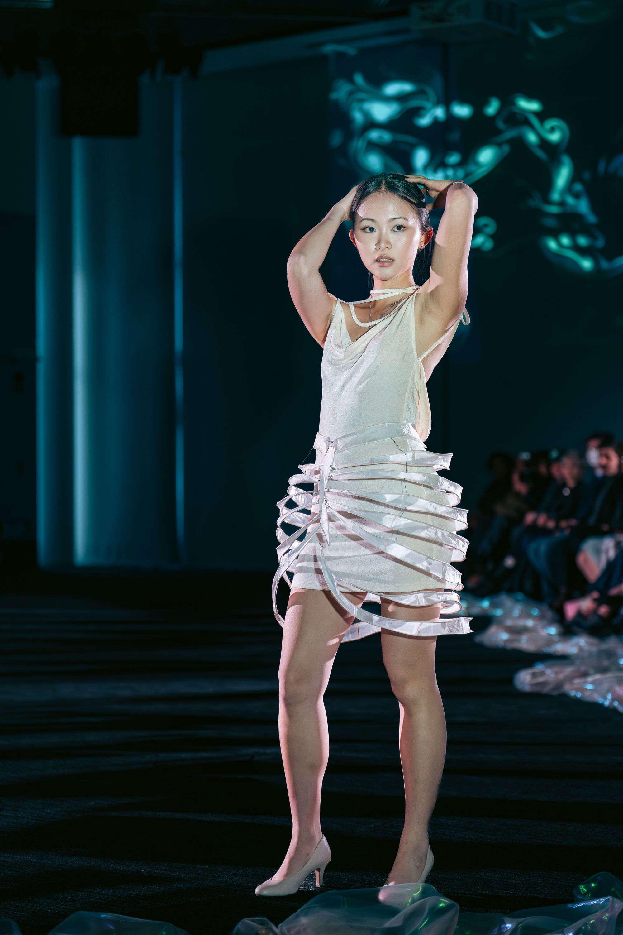 Model faces forward on the runway wearing a cream mini dress with a soft cowl neckline and a sculptural skirt made of translucent, bone-like arcs. Her hands are raised behind her head in a dynamic pose.